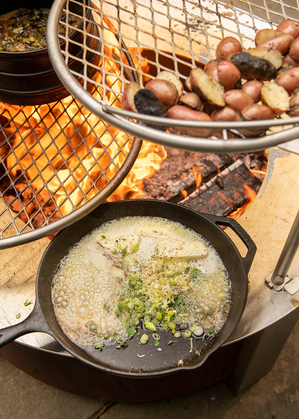 Three dishes being cooked on and above a Breeo fire pit: potatoes, shallots in a skillet, and a pot of stew